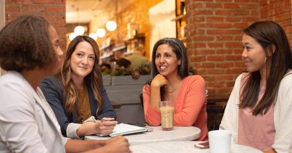 Four women with diverse cultural backgrounds in a business meeting at the table drinking coffee.